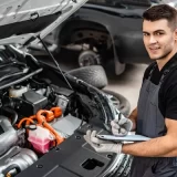 Handsome mechanic writing on clipboard while inspecting car engine compartment